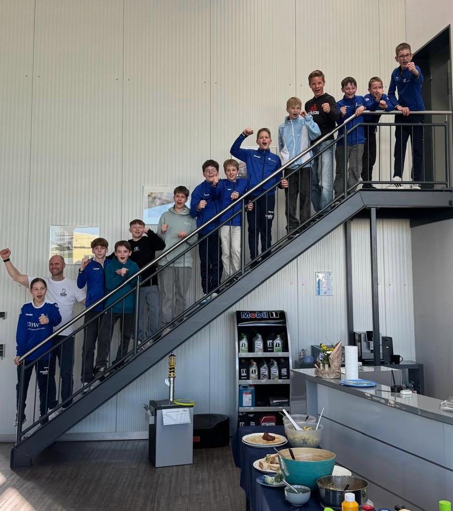 Gruppe von Jugendlichen in blauen Sportjacken, die auf einer Treppe stehen und jubeln, während ein Erwachsener mit ihnen posiert. Im Vordergrund ist ein Tisch mit verschiedenen Speisen und Getränken zu sehen.