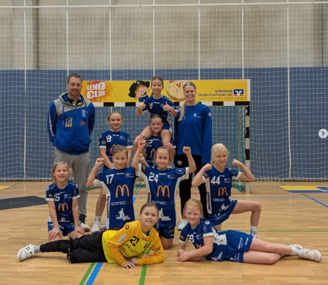 Mädchen-Handballmannschaft in blauen Trikots posiert lächelnd in einer Sporthalle, umgeben von einem Handballtor und Sponsorenbanner. Der Trainer steht hinter der Gruppe.