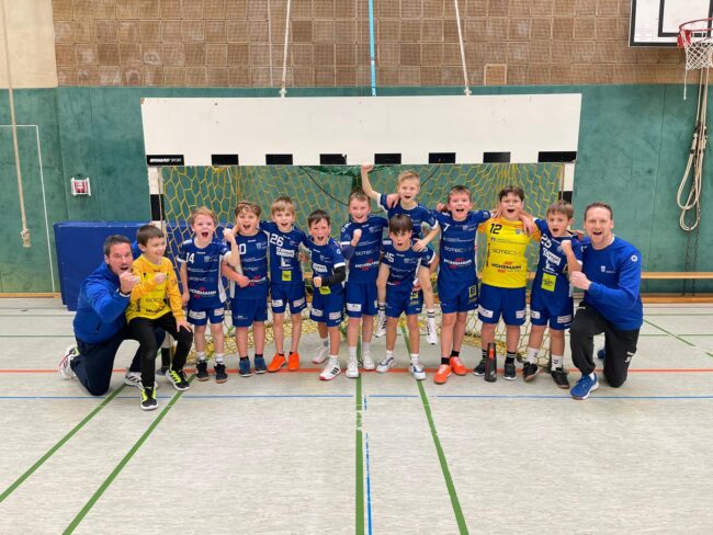 Eine Gruppe von jungen Handballspielern in blauen Trikots feiert mit ihren Trainern nach einem Spiel. Sie stehen vor dem Tor und zeigen ihre Freude über den Sieg. Die Halle ist gut beleuchtet, mit einem Basketballkorb im Hintergrund.