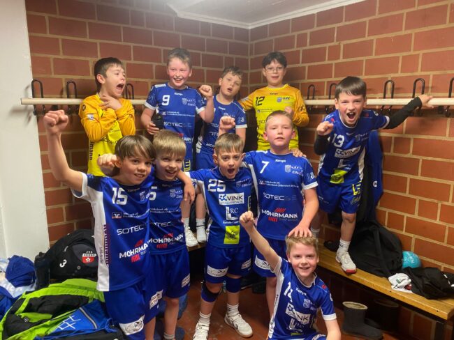 Junge Handballmannschaft in blauen Trikots feiert im Umkleideraum, mit fröhlichen Gesichtern und erhobenen Fäusten, nach einem erfolgreichen Spiel.