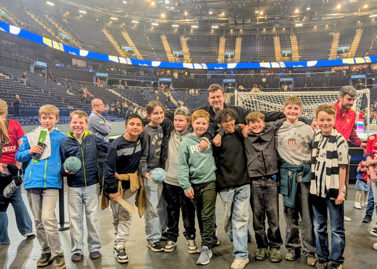 Gruppe von Kindern, die in einer Sportarena stehen, mit einem Trainer oder Betreuer, während sie Handbälle halten. Die Atmosphäre ist lebhaft, mit vielen Zuschauern im Hintergrund und einem Fußballtor sichtbar.